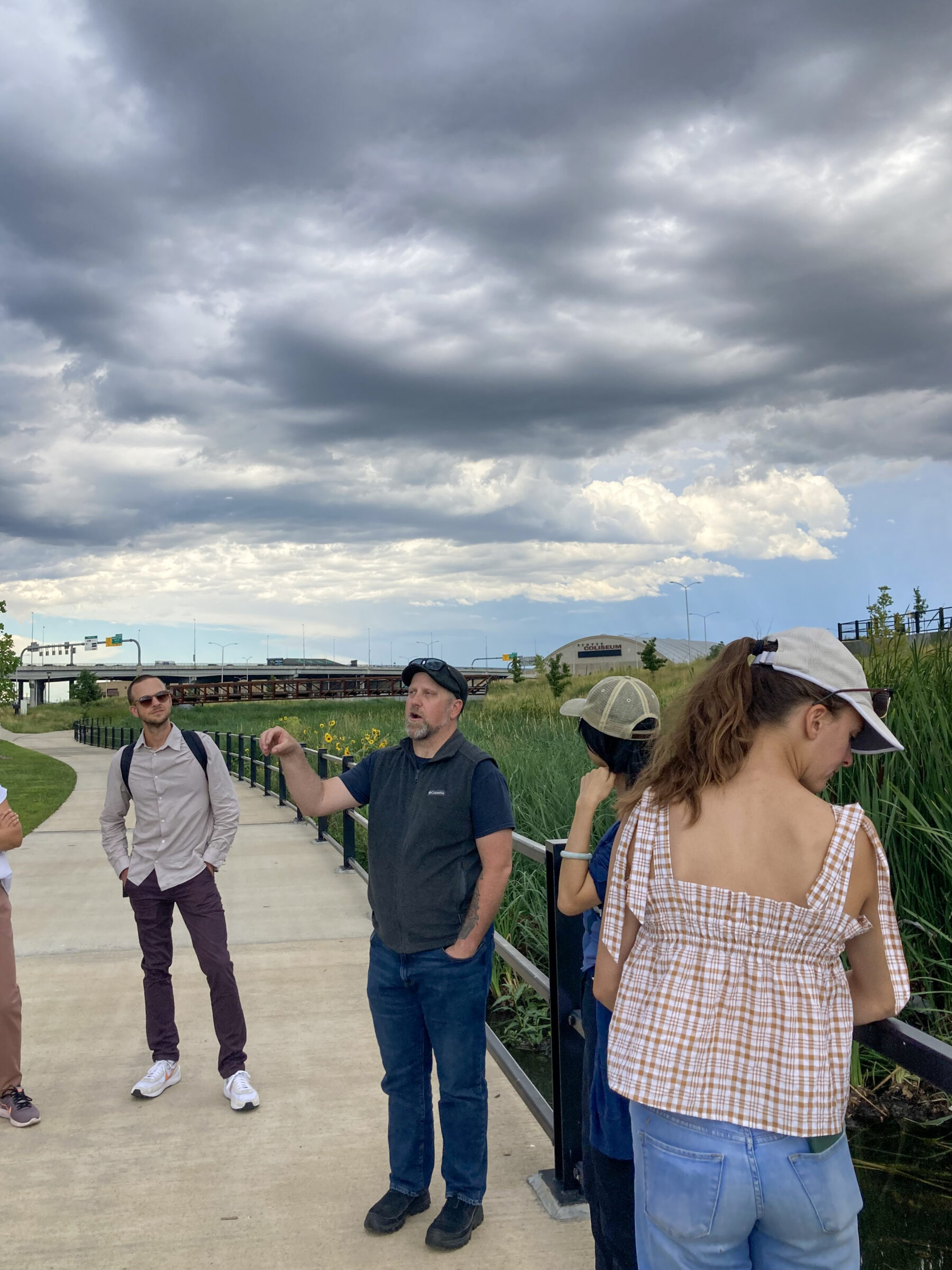 man standing in tall grass addressing small group under cloudy sky