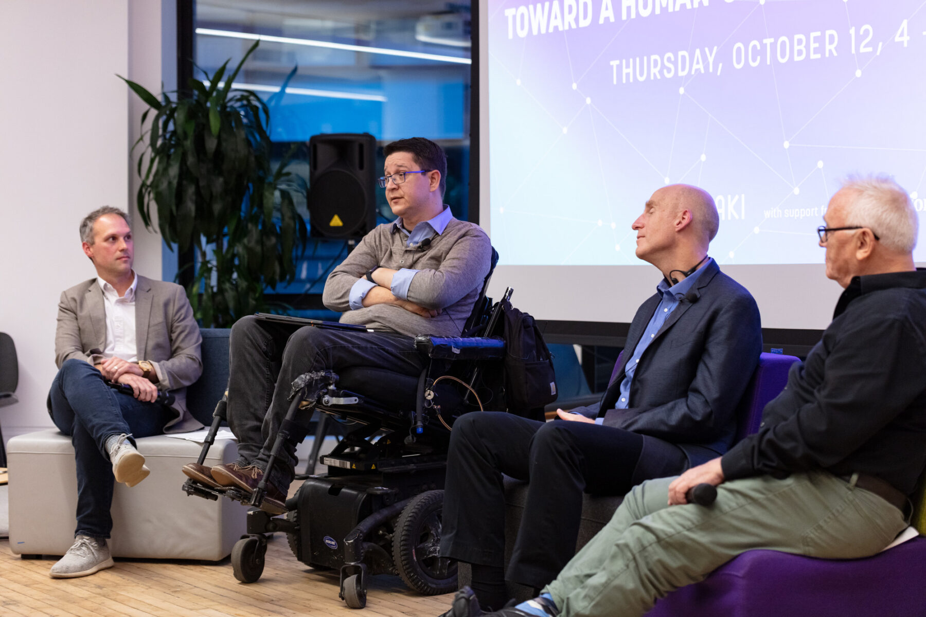 four men sit in front of a projector screen. the three men are looking at the one man speaking, who uses a motorized wheelchair