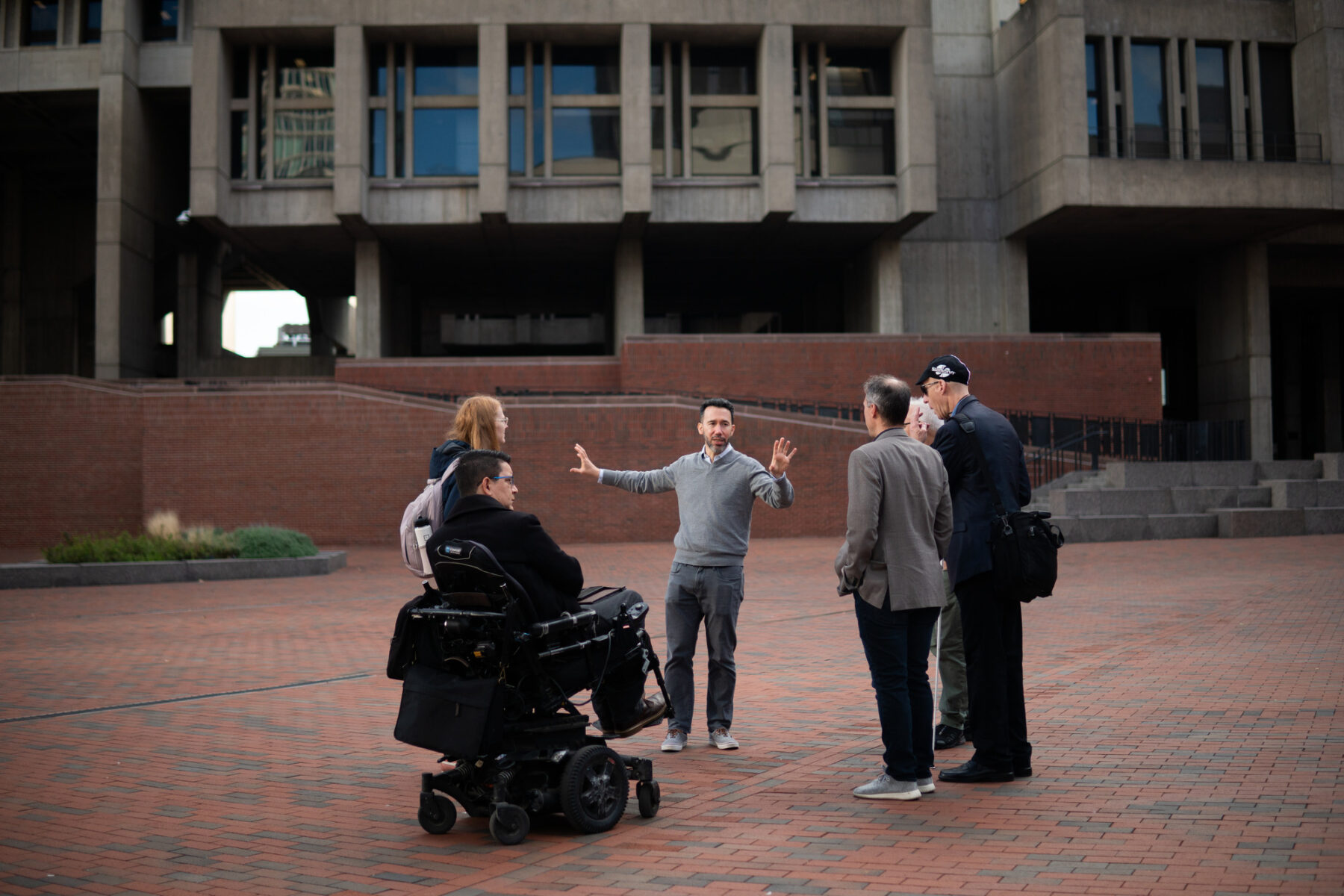 Six people assembled on Boston City Hall Plaza. One person gestures with his arms open towards the space around them