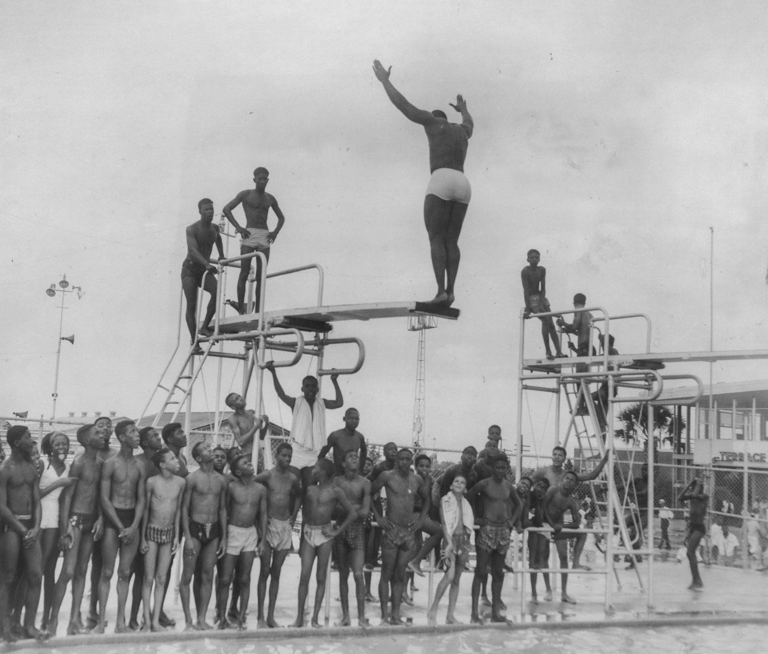 black and white photograph of residents gathered around diving board on beach front