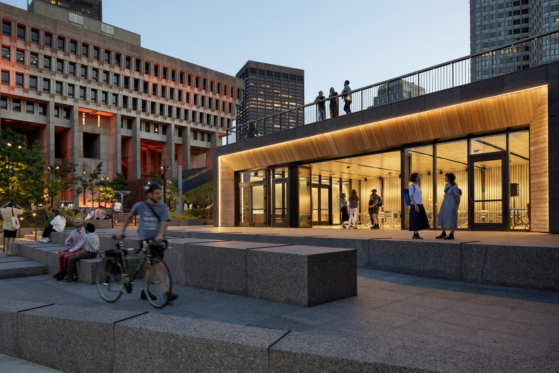 photograph of cyclist walking bike up ramp to venue space shot at dusk, with interior of venue lit up and City Hall plaza in background