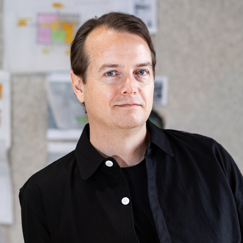 Portrait headshot of Ian Dickenson with pin up wall in studio in background