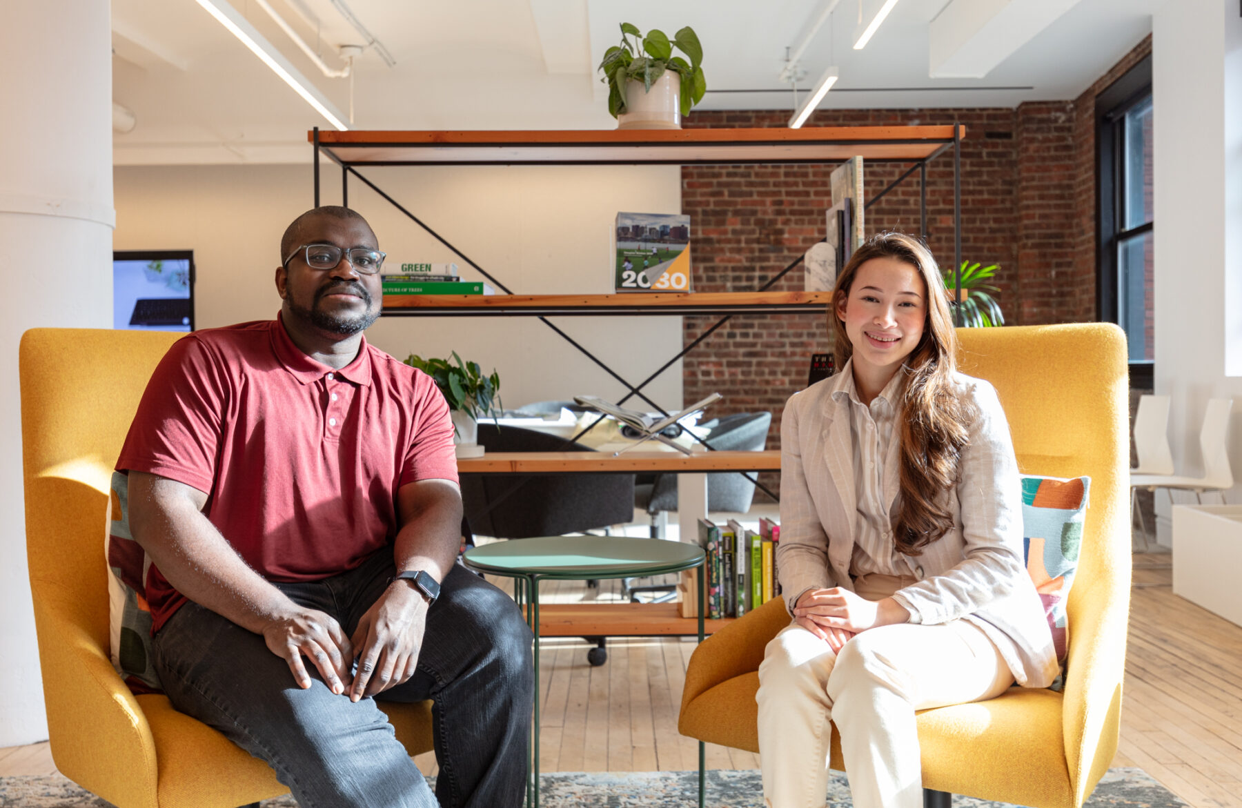 Two people sitting on yellow chairs, smiling, with a wooden bookshelf behind them