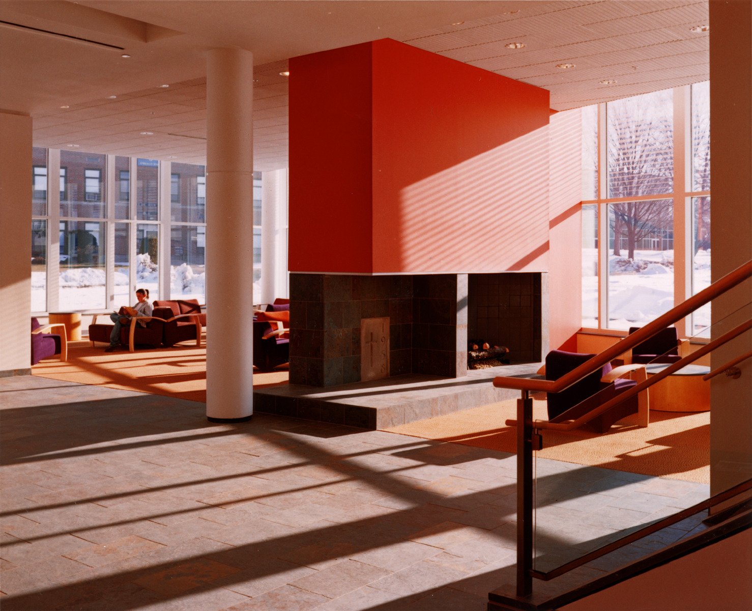 Color photo of a campus center interior. Red fireplace centers an expansive lounge. Outside, a snowy landscape.