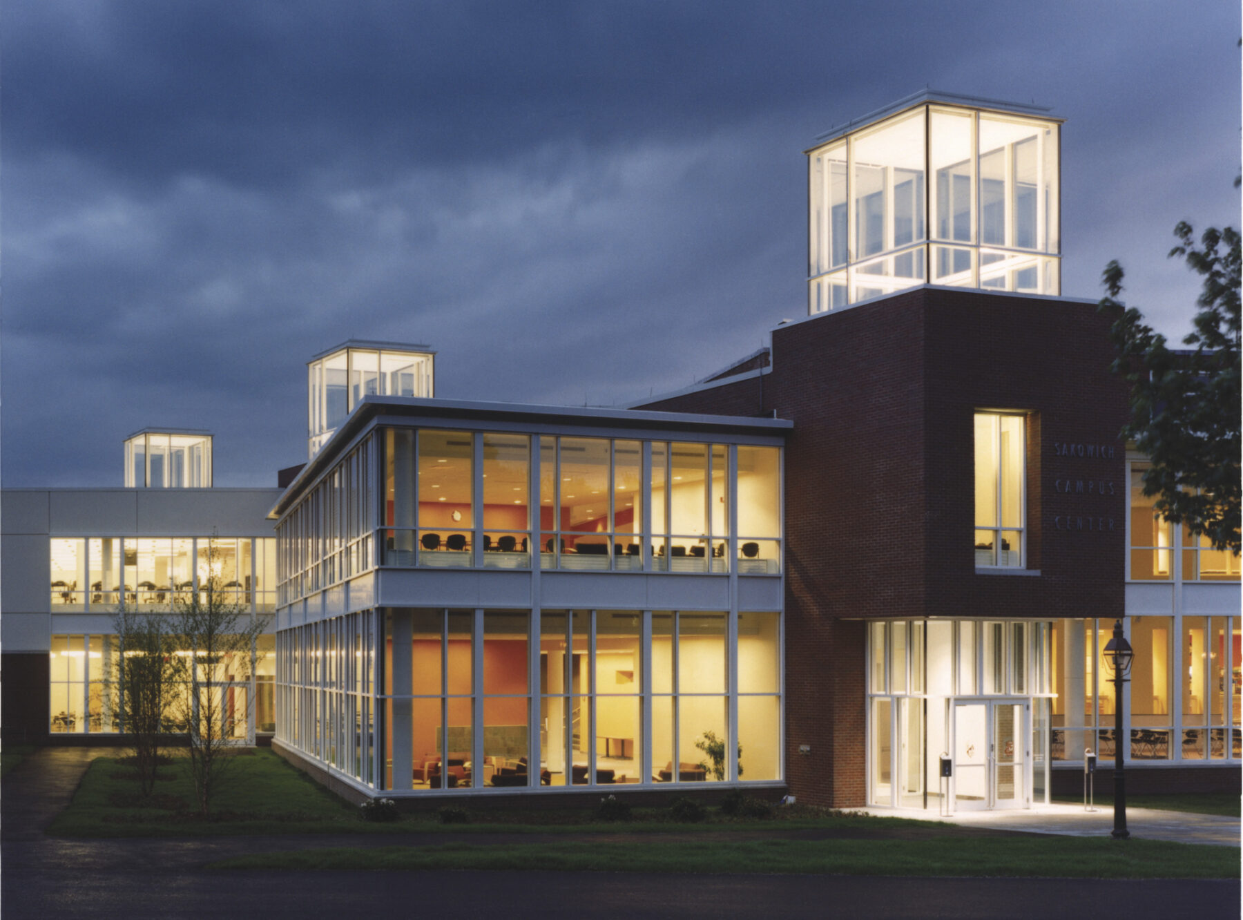 Color photo of a two level campus center, exterior. Three lit glass volumes poke up above the main massing