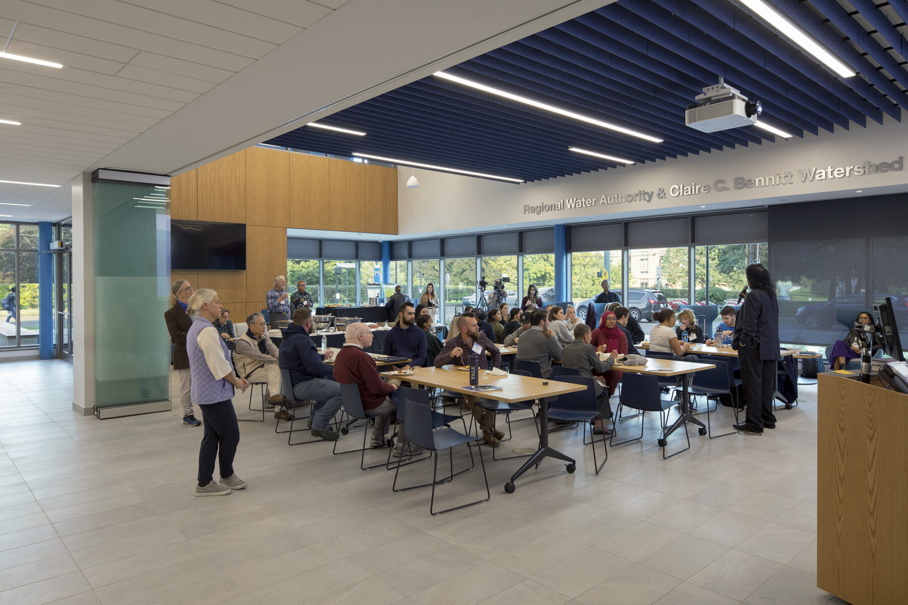 Photo of ground floor level community room with people gathered at tables and speaker addressing the group