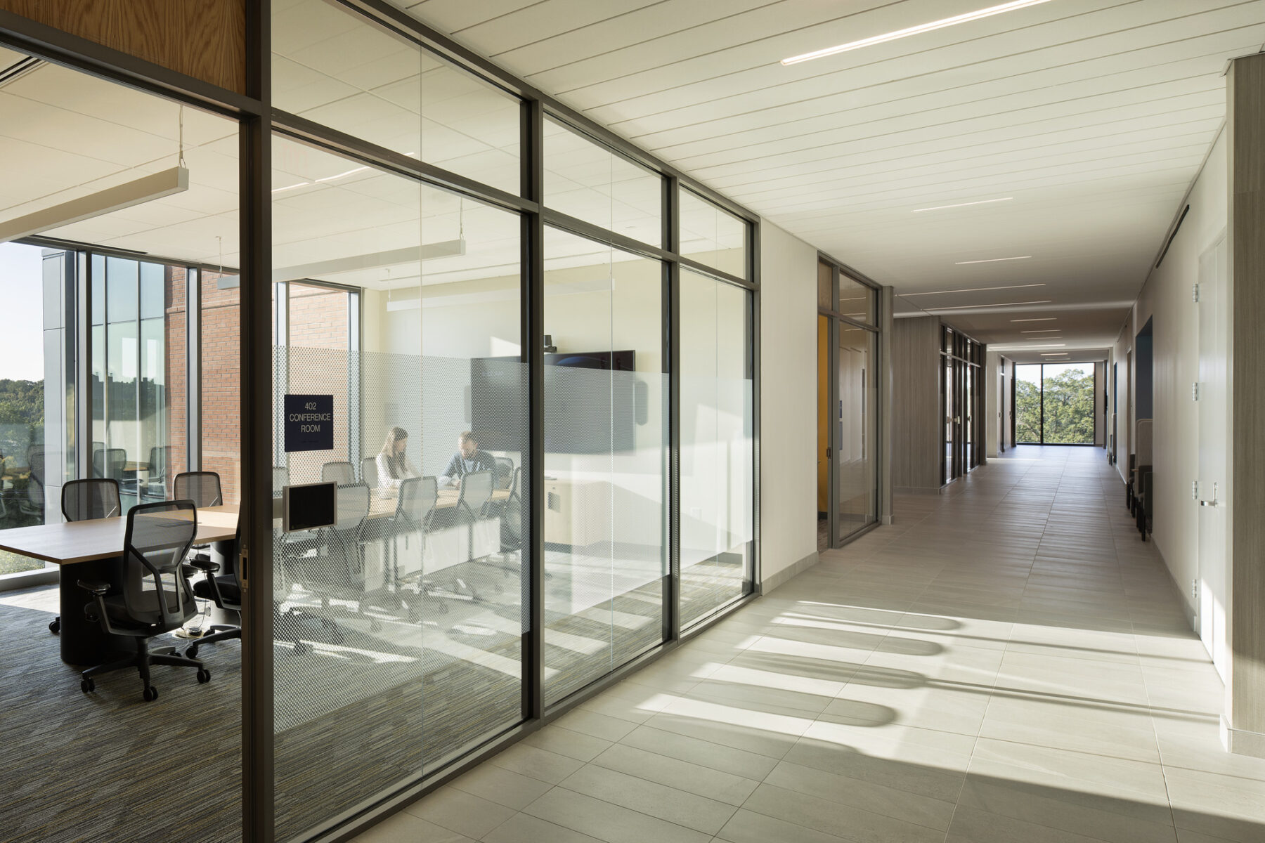 Photo viewing fourth floor hallway outside of meeting room with full height windows and daylight streaming through deep into the building interior