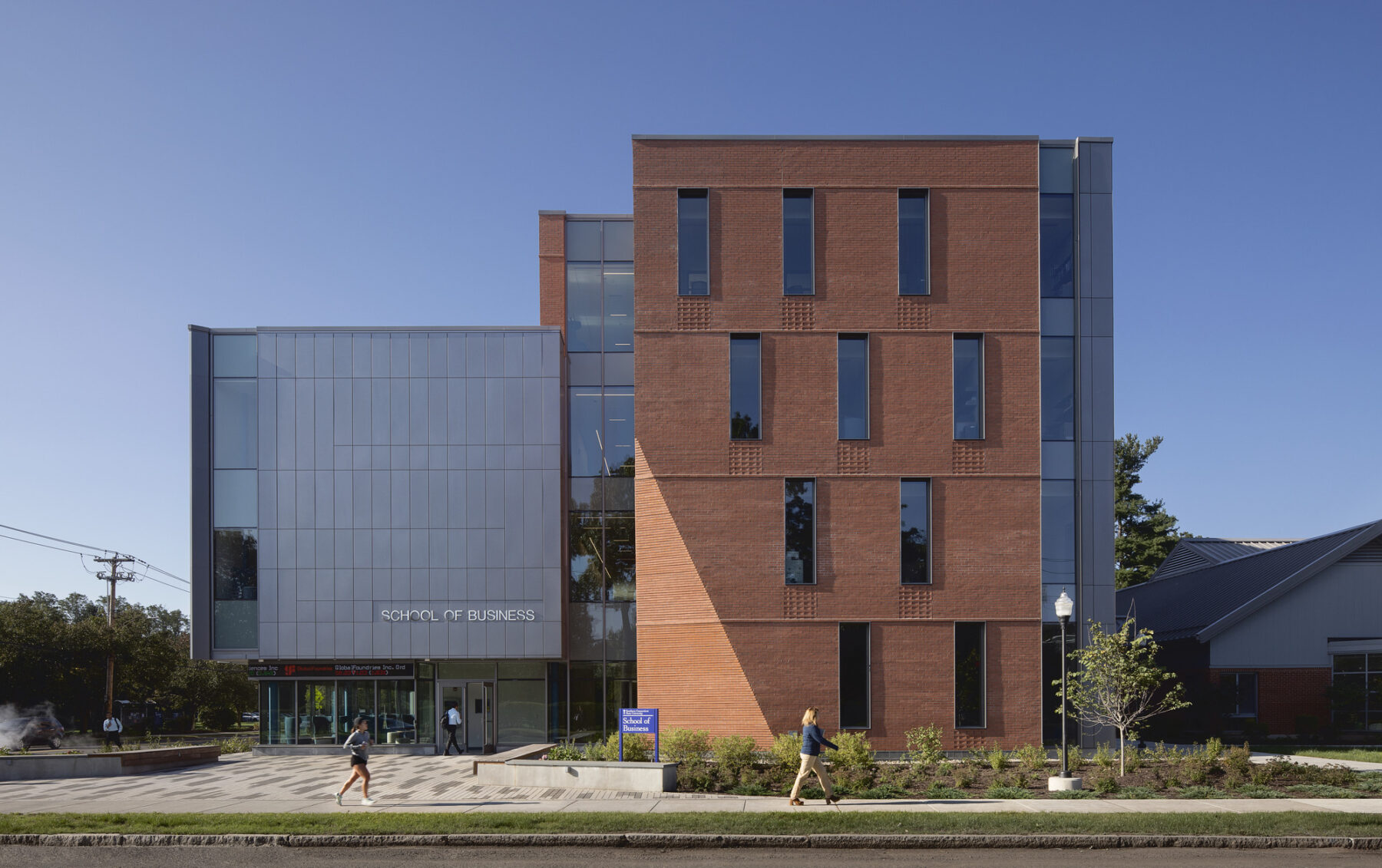 Photo of building exterior with direct elevational view of the entry with sunlight hitting the chamfered brick triangle detail and pedestrians walking in foreground