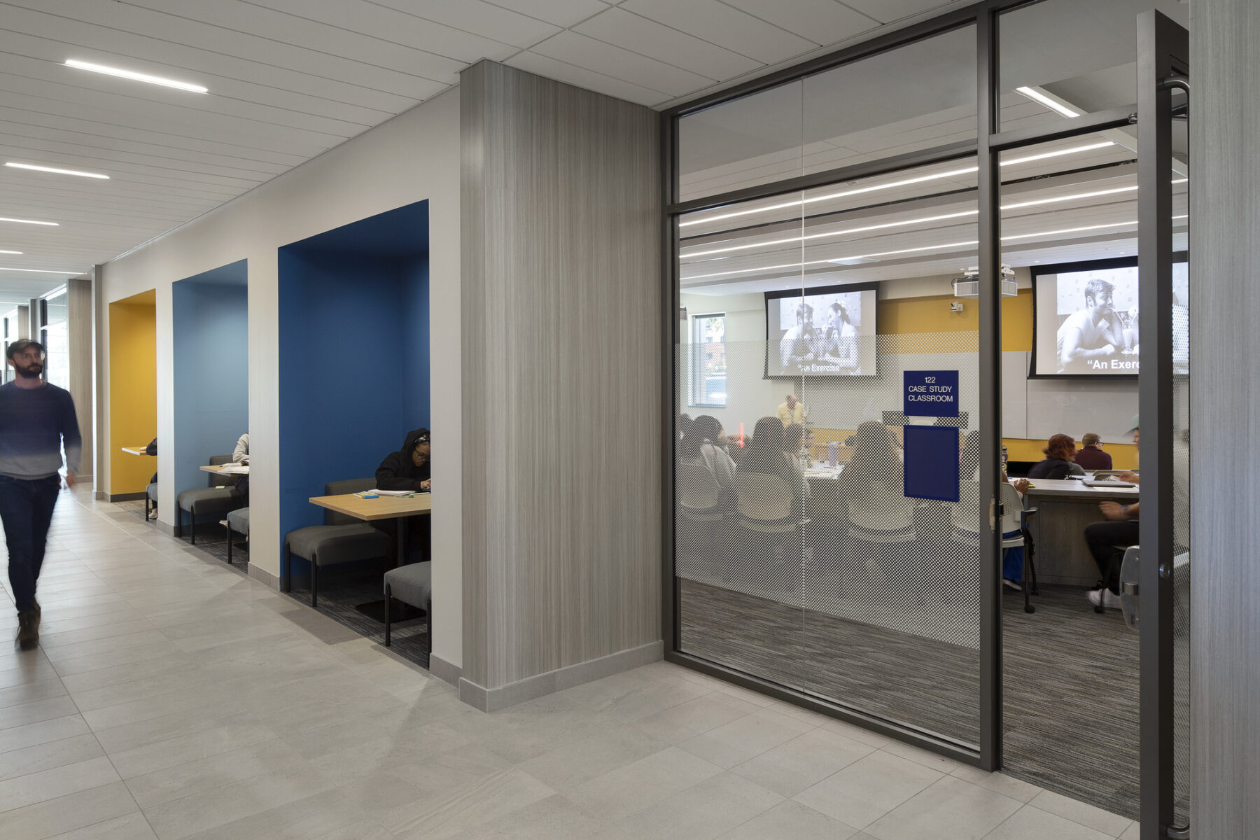 Photo looking down the hallway with view into classroom and colorful nooks with tables and students engaged in casual study sessions