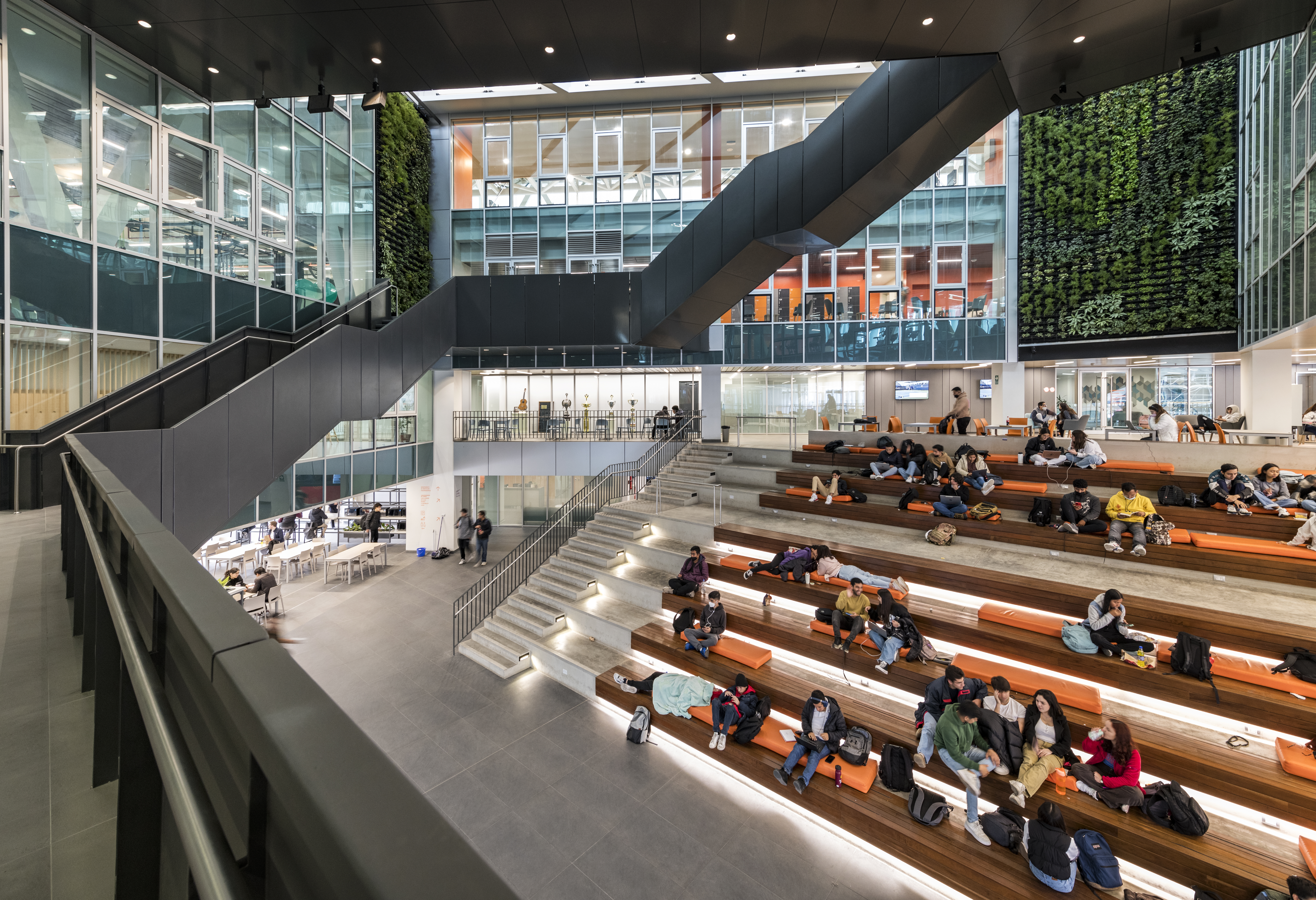 Color photo. Students sit on terraced seating in wide atrium in front of green wall. stairs wind around the perimeter of atrium. on upper levels, exercise rooms are visible