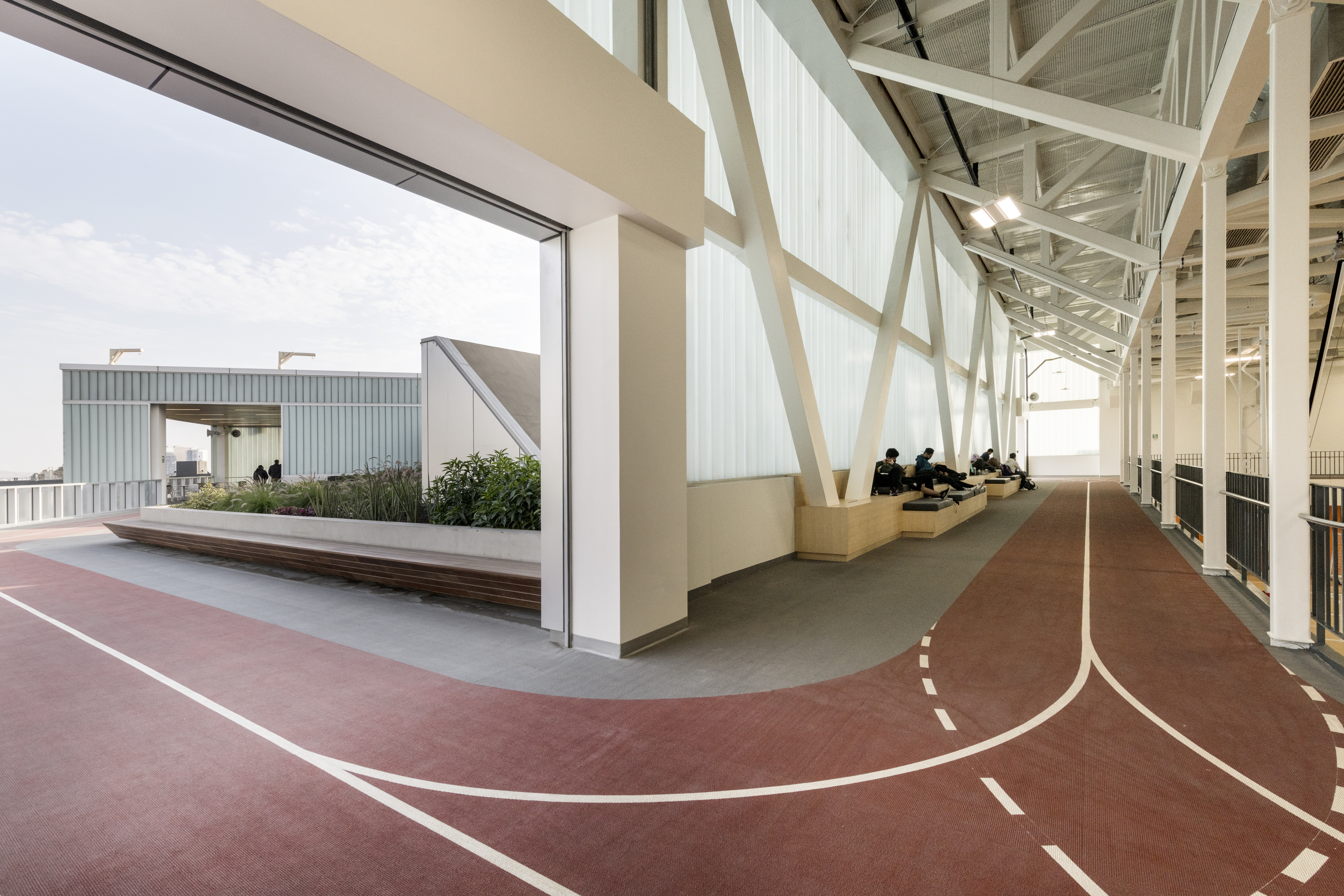 Color photo. On the building roof, a track weaves between covered spaces and the open sky. Students walk the track and study on adjacent benches.