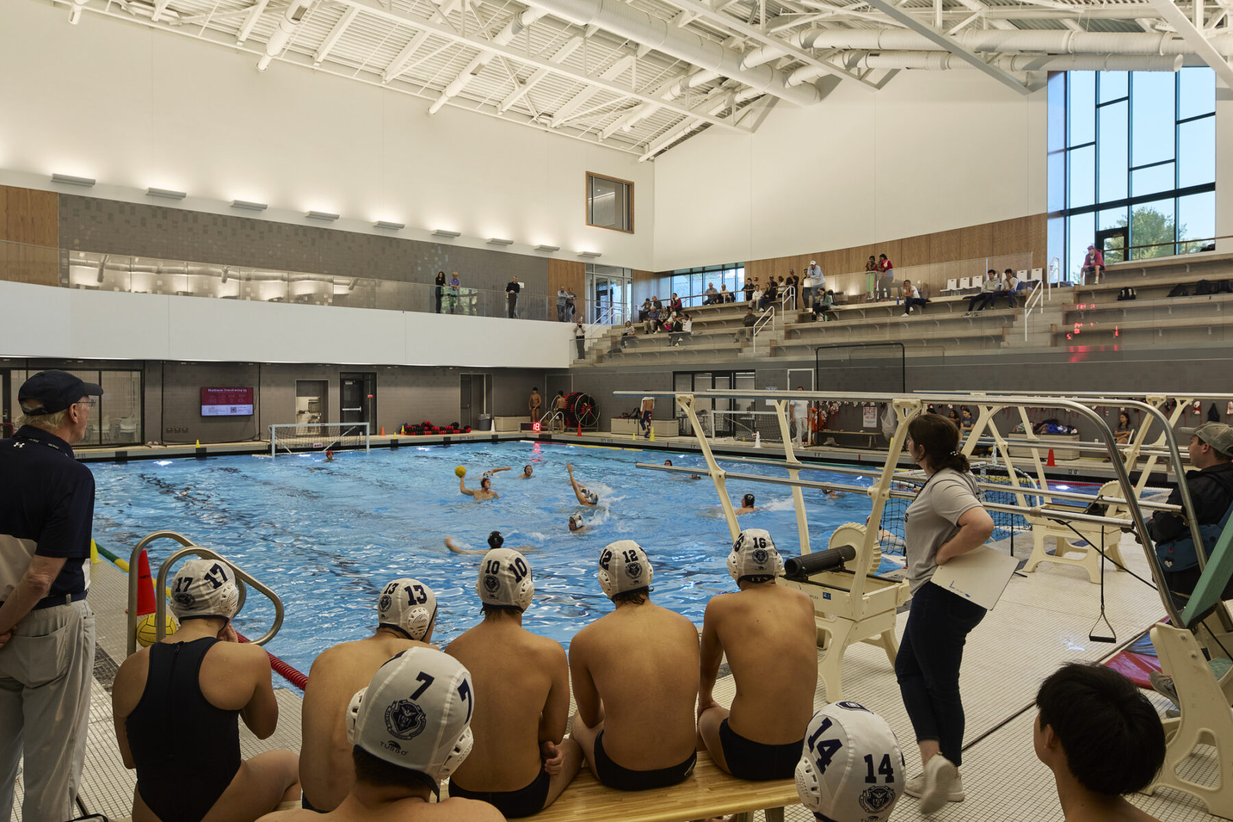 Inside the pool area, with a view from behind the water polo team facing the pool and the upper level spectator bleacher seating and the curved ceiling framing the space from above.
