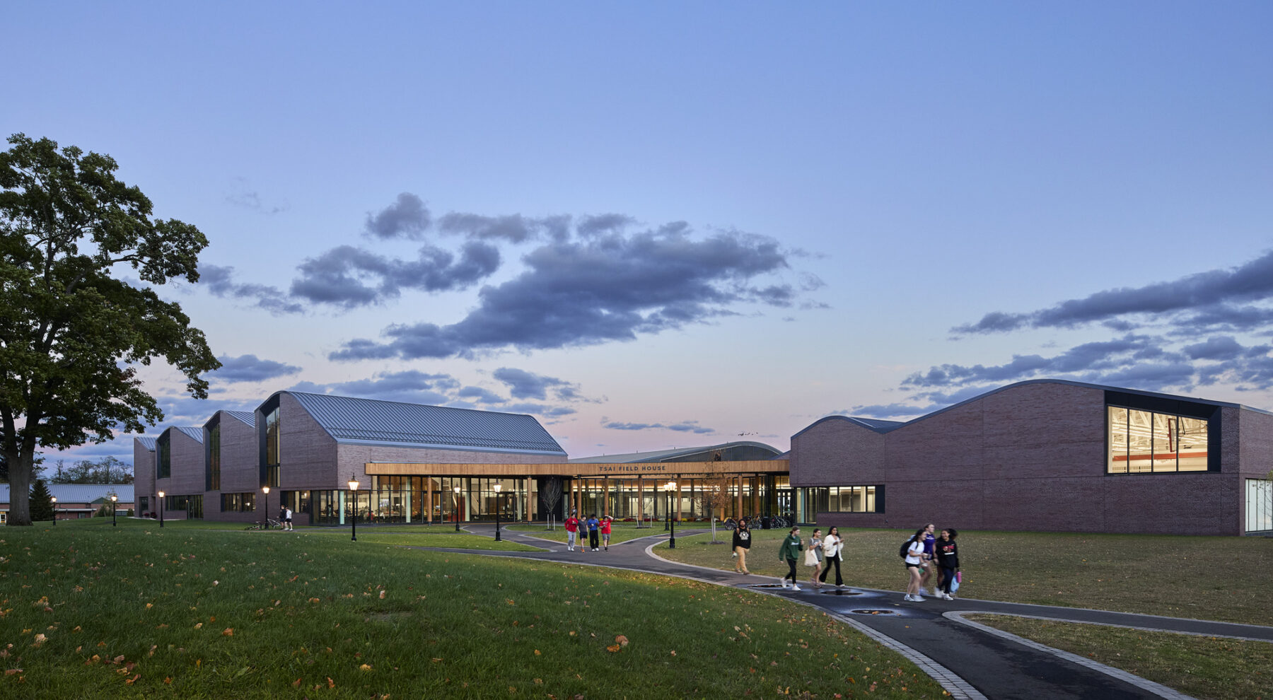Exterior at dusk facing the Tsai Commons and Field House from across the campus with circulation paths leading to the glowing lights inside