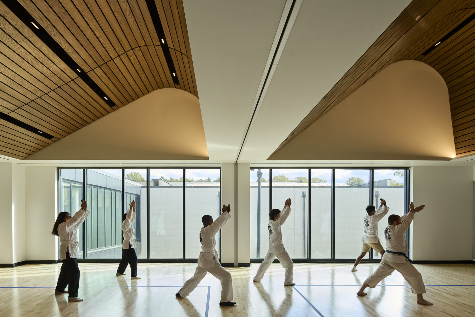 A karate class inside the multipurpose fitness room, with floor to ceiling windows and a wooden ceiling with two vaulted, curved profiles above.