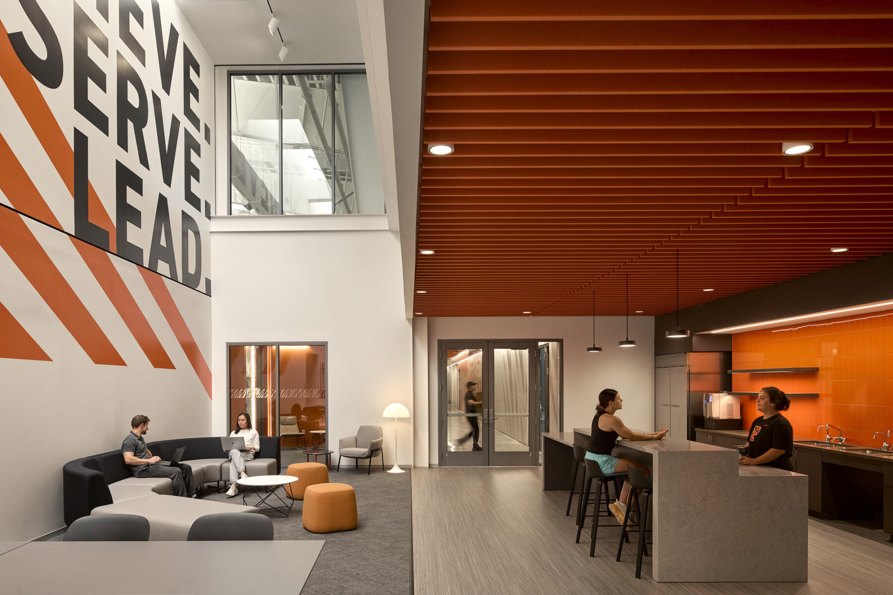 photo of interior student lounge with flexible furniture, a kitchen with counter and bar stools, and views through glass panels into indoor tennis arena