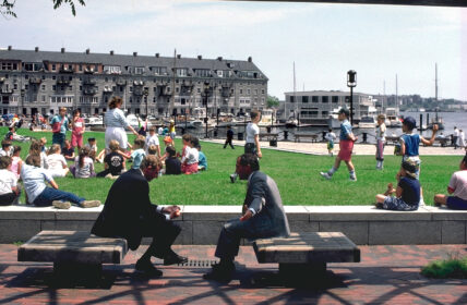 Old photo of 2 men sitting on park benches with view of the lawn and wharf in distance