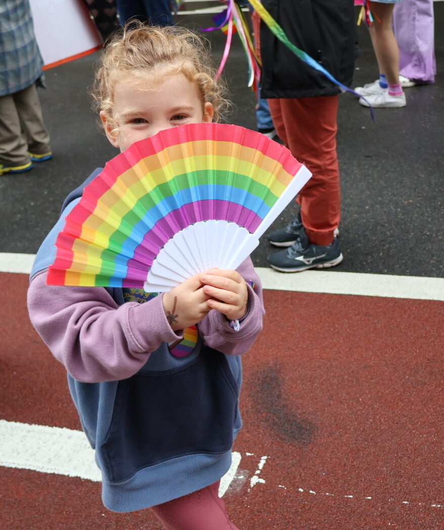 A young girl holds a rainbow fan in front of her face and poses for a photo