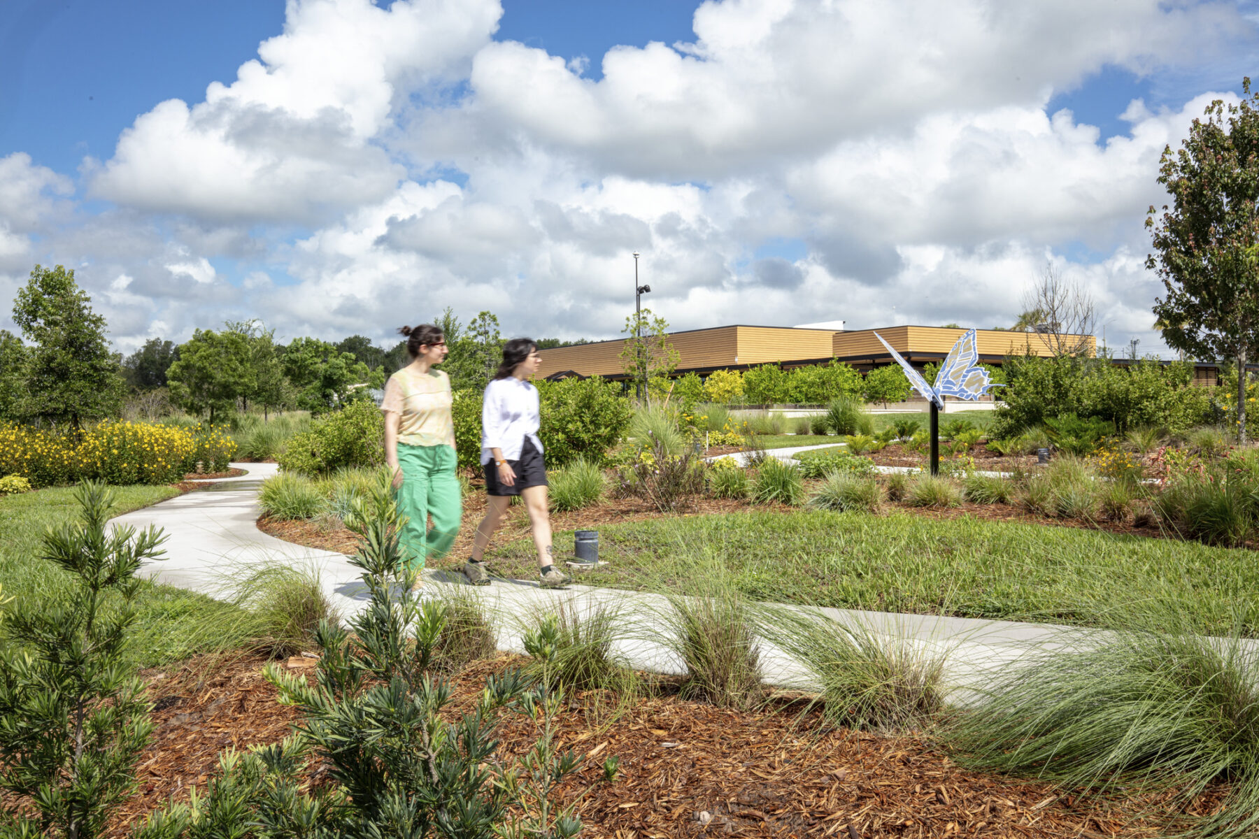 two people walk on a paved path surrounded by grass and plants