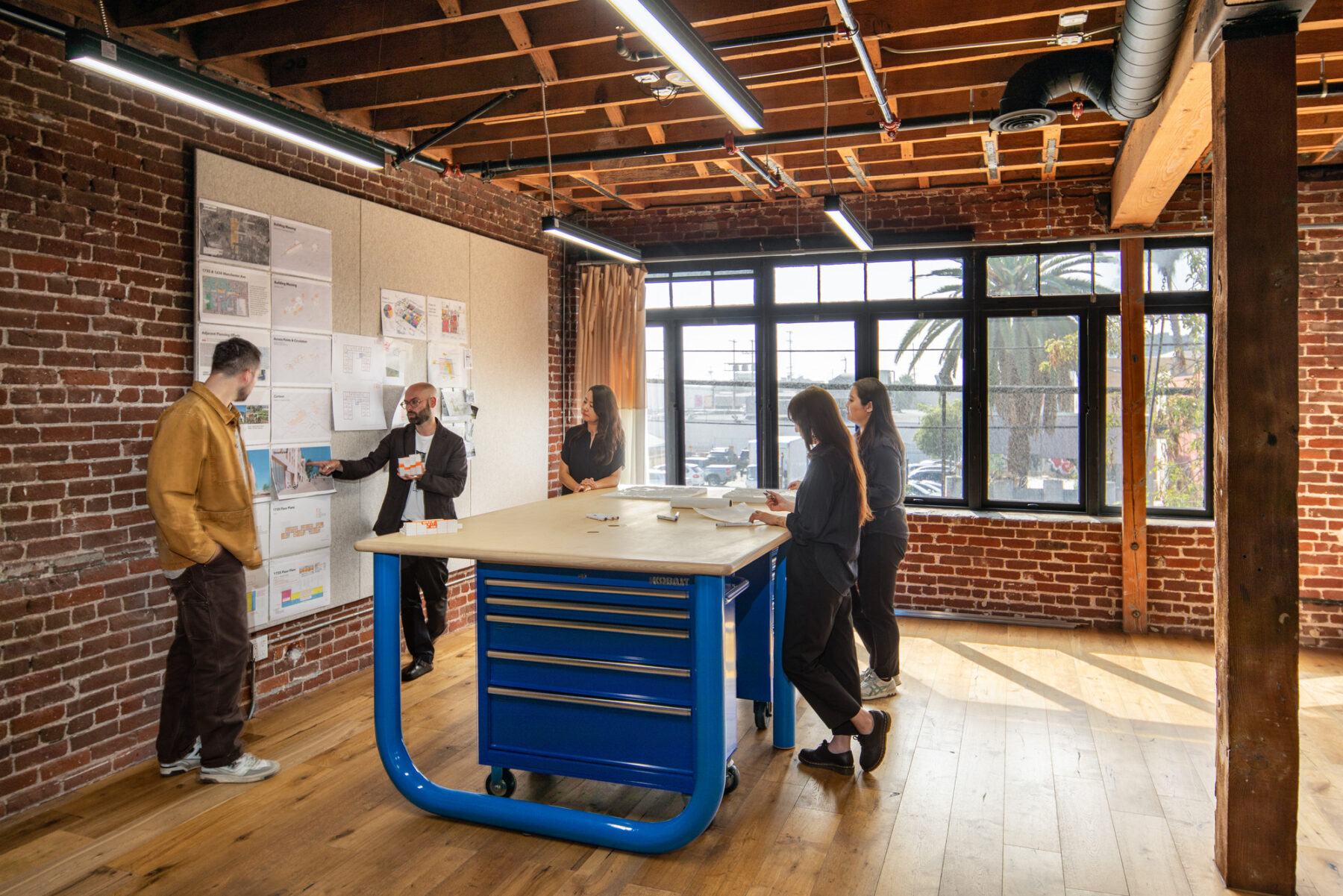 a photo of a group of people gathered around a table, looking at materials pinned up on a wall