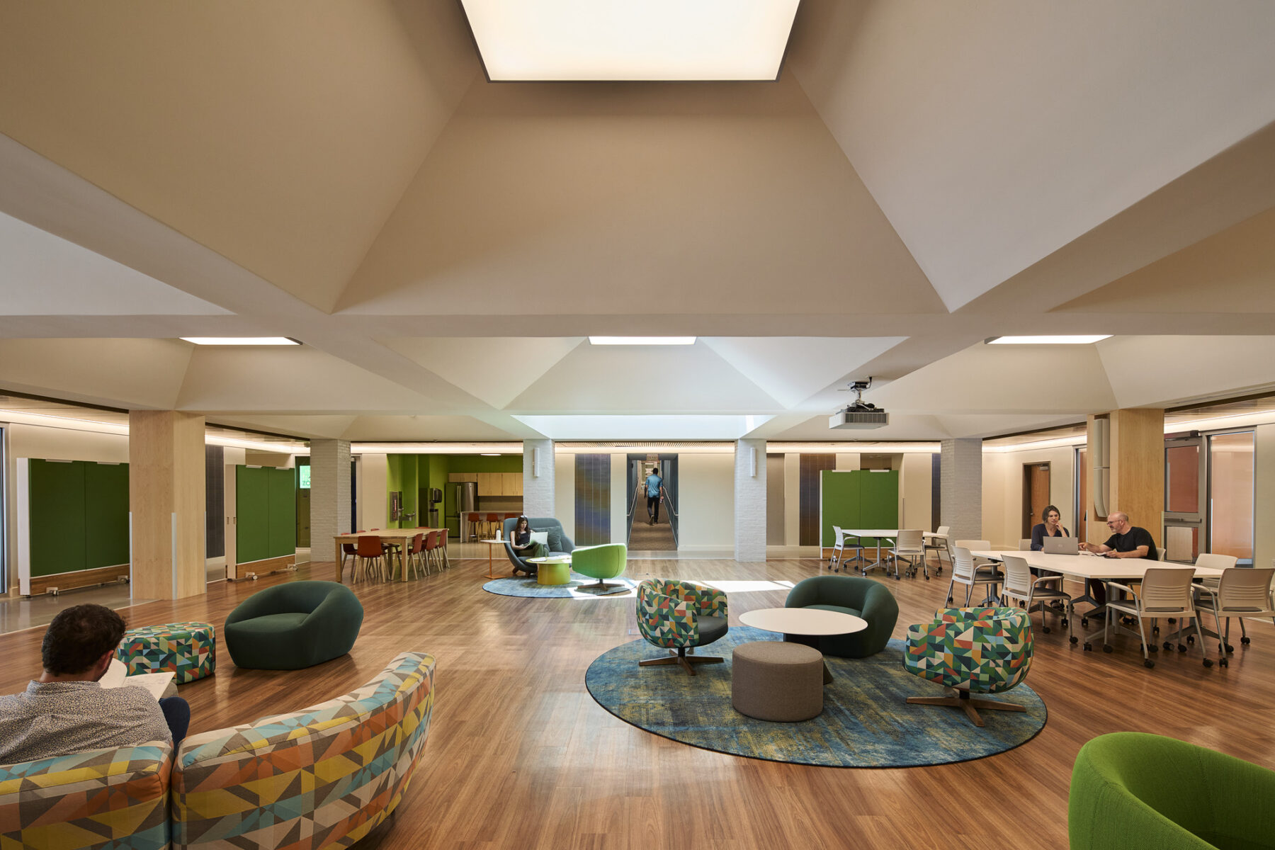 Interior photo of Commons space with lounge furniture, work table, and view of ramp hallway leading to dorm