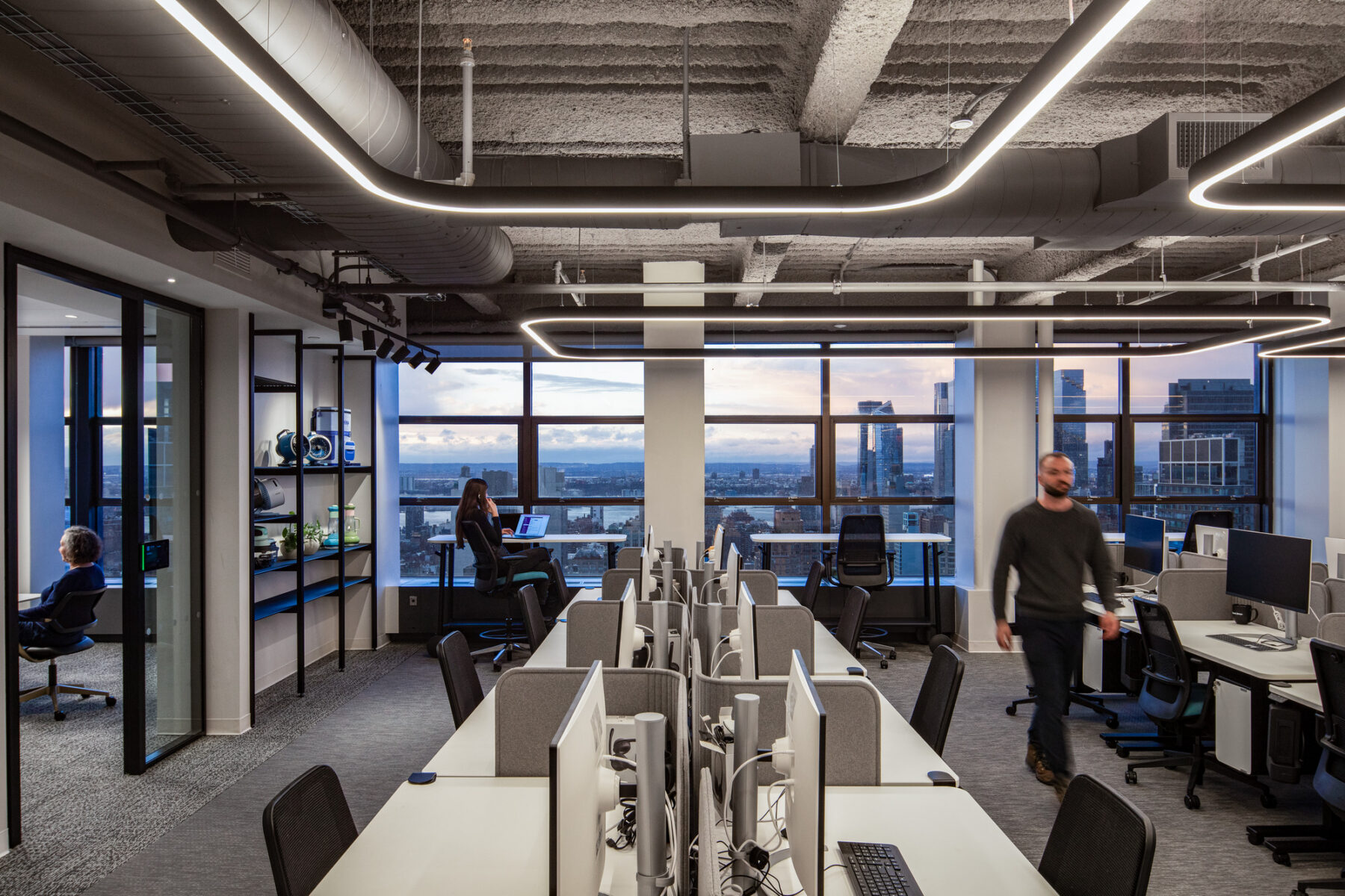 photograph of an open desk area in corporate office setting with one person walking and another taking a zoom call in the background