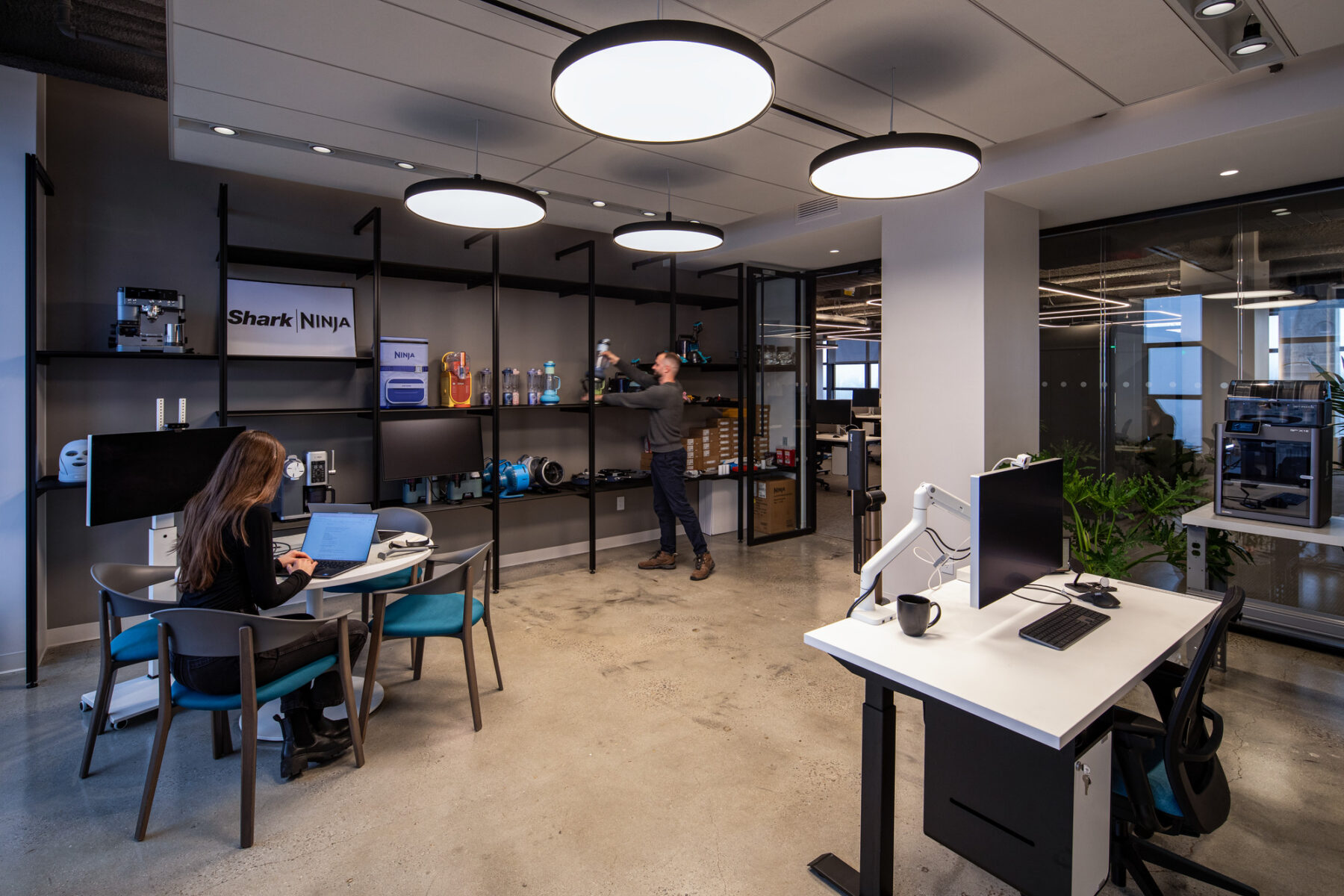 photograph of corporate setting showing two people in a private office with a desk, bookshelf, meeting table and circular light fixtures in the ceiling.
