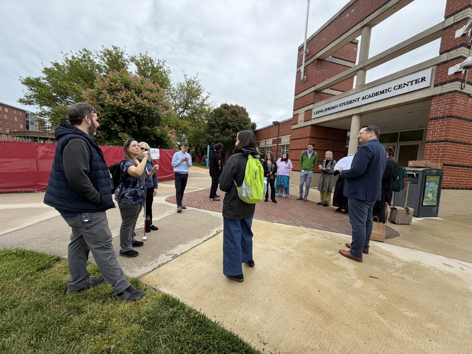 A group of 15 adults stand in a circle outside. They are next to a university Student Center looking at a woman using American Sign Language.